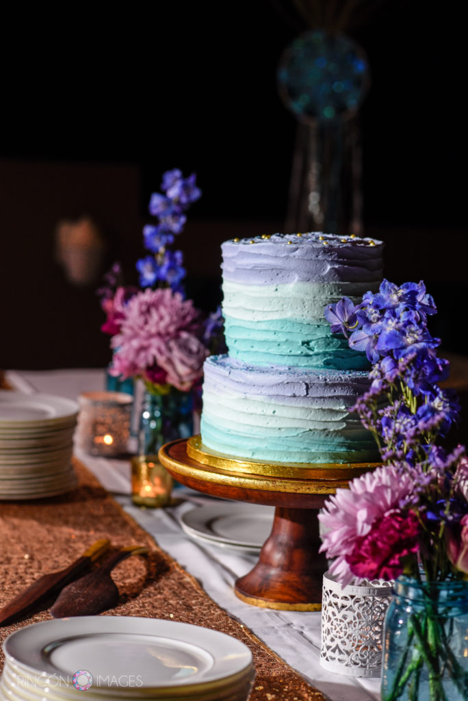 Purple and teal frosted wedding cake with purple and pink flowers on a table.