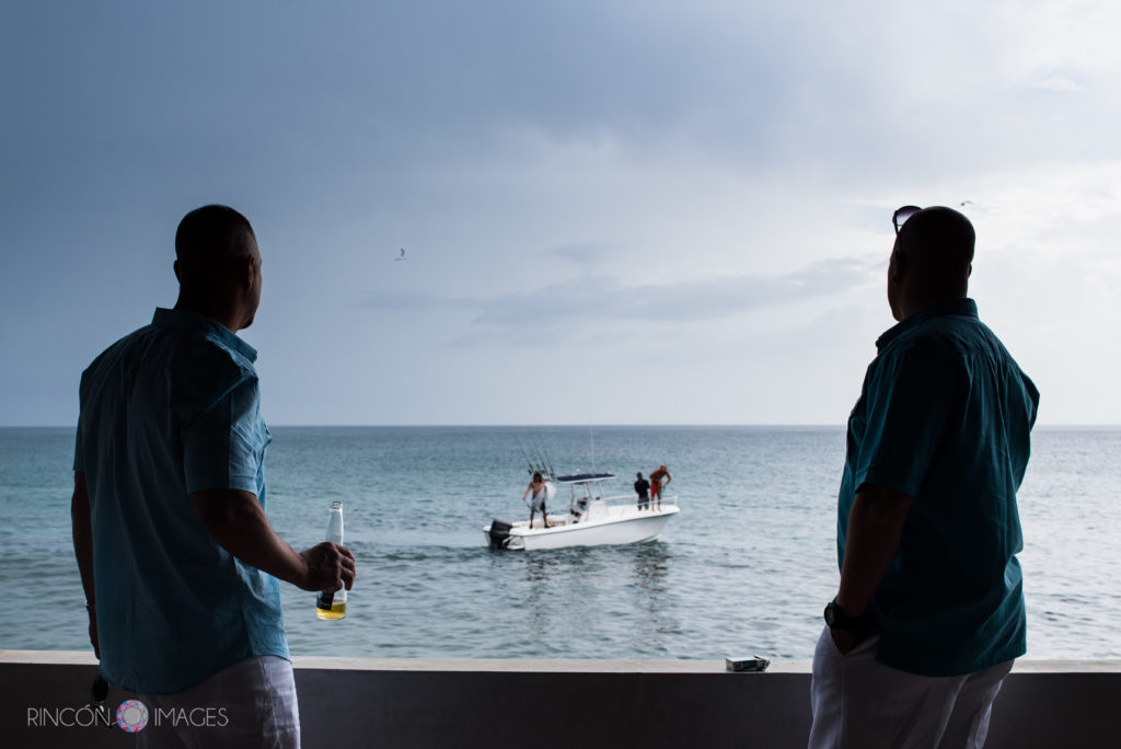 Two groomsmen wearing matching teal shirts look off the porch ledge at a fishing boat in the ocean.